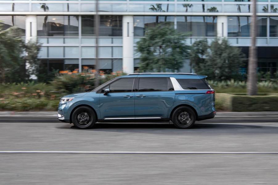 The side profile of a blue Kia Carnival driving on the street in front of a building.