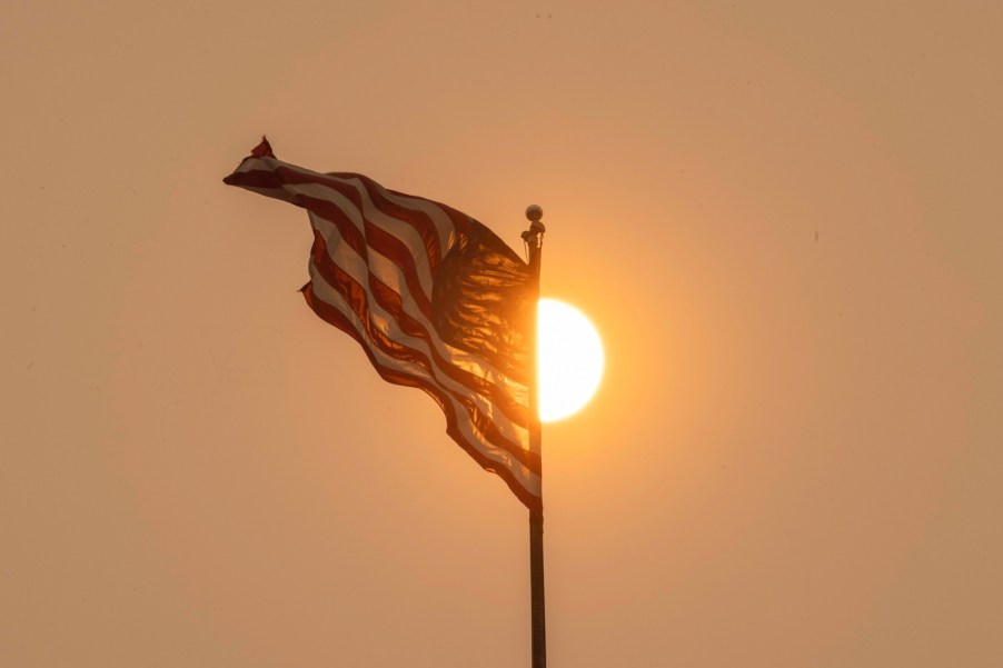 A U.S.A. flag waves in front of a hazy, setting sun like the flags in front of auto plants in the U.S.