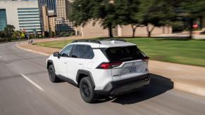 A rear side shot of a white 2022 Toyota RAV4 TRD Off-Road compact SUV model driving through a city