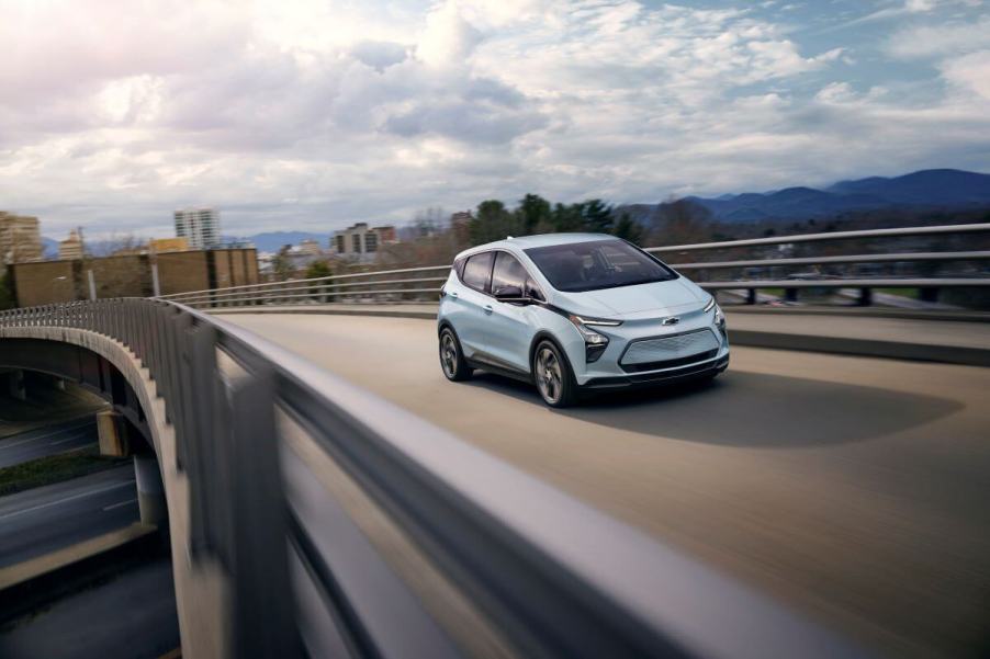 A light blue 2023 Chevy Bolt EV hatchback model driving on a highway overpass