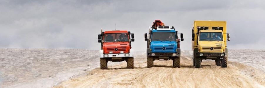 A line of Mercedes-Benz Unimog models in red, blue, and yellow driving down a road in a desert