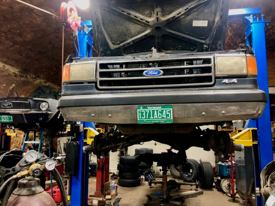 The grill of a black 1988 Ford F-150 XLT Lariat square body classic truck that is on a garage lift with its tires off and hood up.