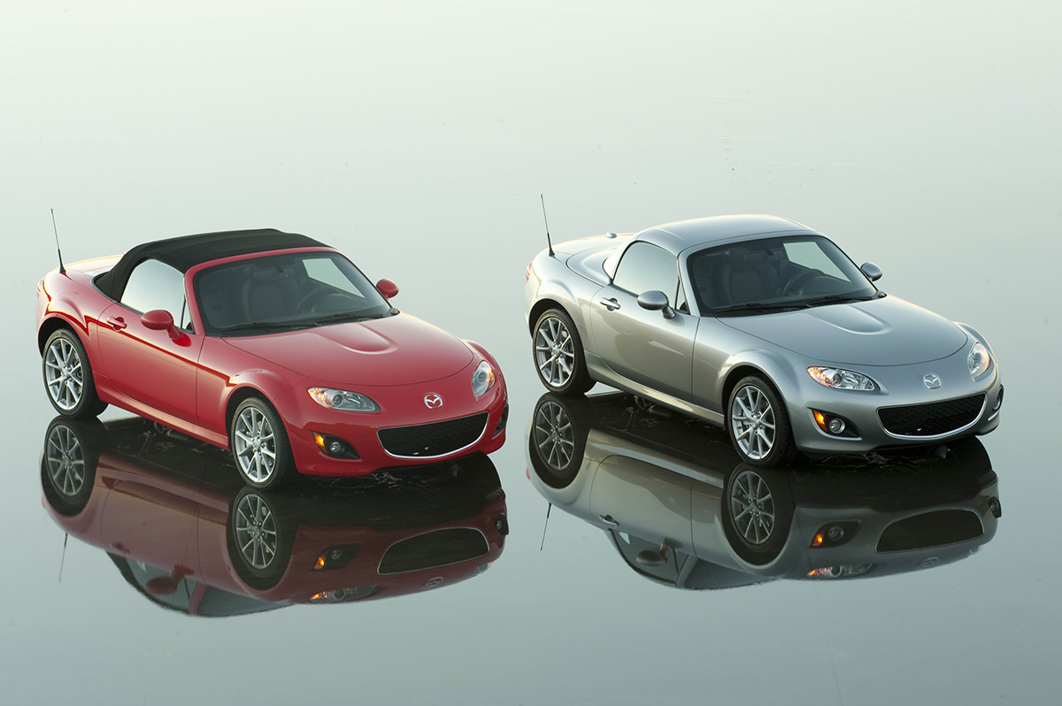 A pair of NC Miata twins sitting on a reflective lakebed surface. One red with a soft top and one silver with a hard top. 