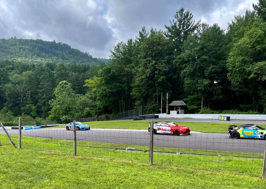 An Aston Martin, BMW, and Porsche GT4 racecars ascend the hill at Lime Rock Park