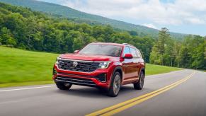 A 2024 Volkswagen Atlas three-row midsize SUV model in Aurora Red Metallic driving out of a green forest