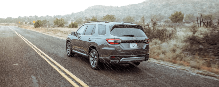 A rear shot of a 2024 Honda Pilot Elite midsize SUV in Modern Gray Metallic driving through desert rain