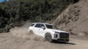 A white 2024 Hyundai Palisade driving on a dusty road. The Hyundai Palisade price is a perk compared to the Infiniti QX60