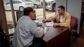 A salesperson and a car buyer discuss a contract over a table at a dealership, used trucks visible in the background.