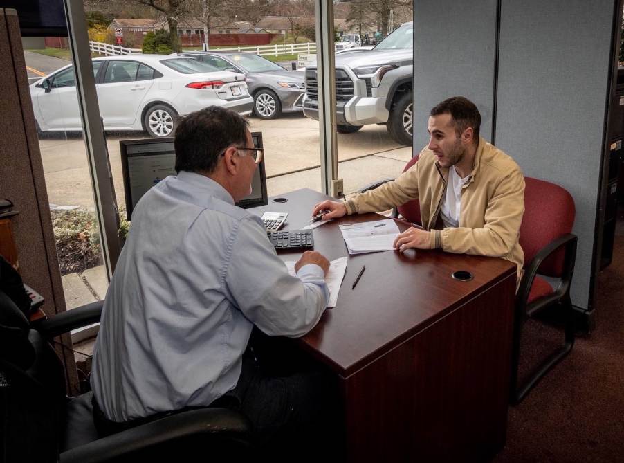 A salesperson and a car buyer discuss a contract over a table at a dealership, used trucks visible in the background.