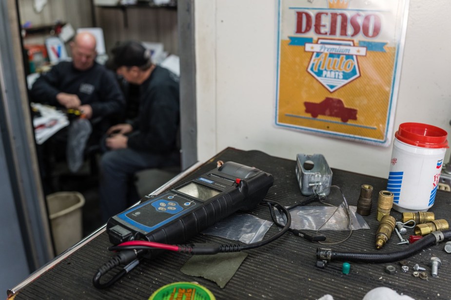 Two mechanics sit in a distant office, a table of tools visible in the foreground.