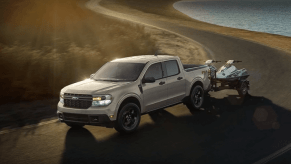 A 2024 Ford Maverick compact pickup truck model towing a pair of ATVs past a lake