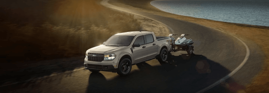 A 2024 Ford Maverick compact pickup truck model towing a pair of ATVs past a lake