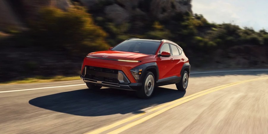 An orange 2024 Hyundai Kona subcompact SUV is driving on the road.