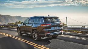 A 2024 Jeep Grand Cherokee L Summit Reserve midsize SUV parked on a coastal highway with a bridge in the distance