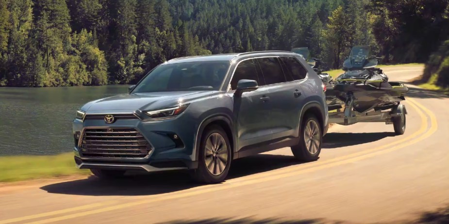 A blue fully loaded 2024 Toyota Grand Highlander Hybrid MAX is driving while towing a jet ski. 