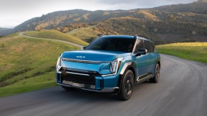 A blue 2024 Kia EV9 midsize electric SUV is driving on the road.