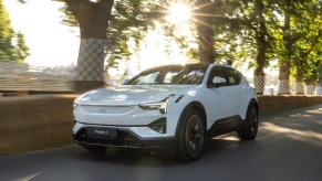 A white Polestar 3 driving on a road in the woods. Compare the Polestar 2 and Polestar 3.