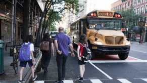 Students board a school bus on a city street in Manhattan New York, a grownup walking behind them.