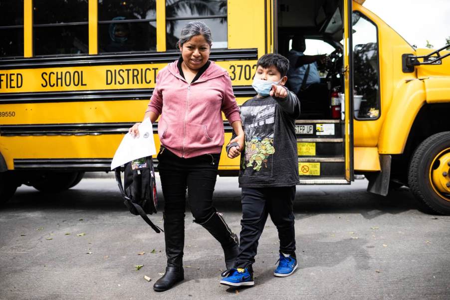 A little boy points while his mother leads him away from a school bus stop.