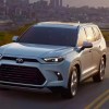 A white 2024 Toyota Gand Highlander Hybrid midsize three-row hybrid SUV is driving on the road.