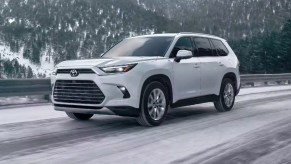 A white 2024 Toyota Grand Highlander midsize three-row SUV is driving on a snowy road.