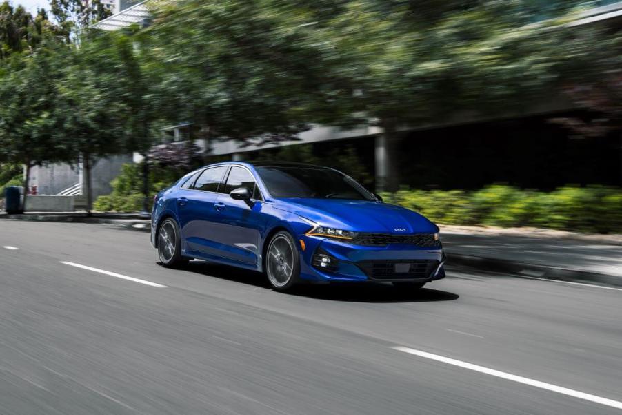 A 2024 Kia K5 midsize sedan model driving through a city past a line of trees