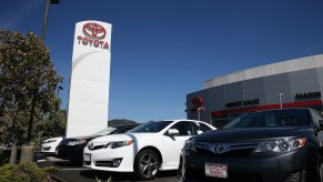 An expansive Toyota dealership lot under the clear blue sky, featuring rows of shiny new and pre-owned vehicles neatly arranged in orderly rows. The showroom building stands prominently at the center, with a sleek glass facade and the Toyota logo prominently displayed. Customers and sales staff can be seen inspecting cars, and colorful Toyota banners and flags flutter in the gentle breeze, adding a vibrant touch to the scene.