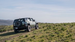 Rear passenger-side shot of the 2024 GMC HUMMER EV SUV in Moonshot Green Matte with Sky Panels removed climbing a grassy off-road hill. GMC Hummer EV sales are on the rise.