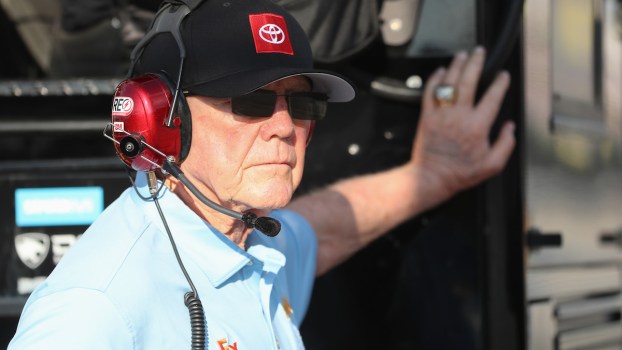 Joe Gibbs Racing is helmed by Joe Gibbs himself, who leans against a wall.