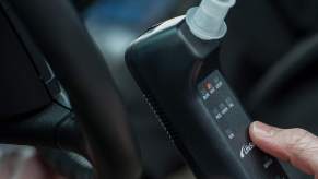 A hand holds a breath alcohol ignition interlock device (IID) next to a car steering wheel