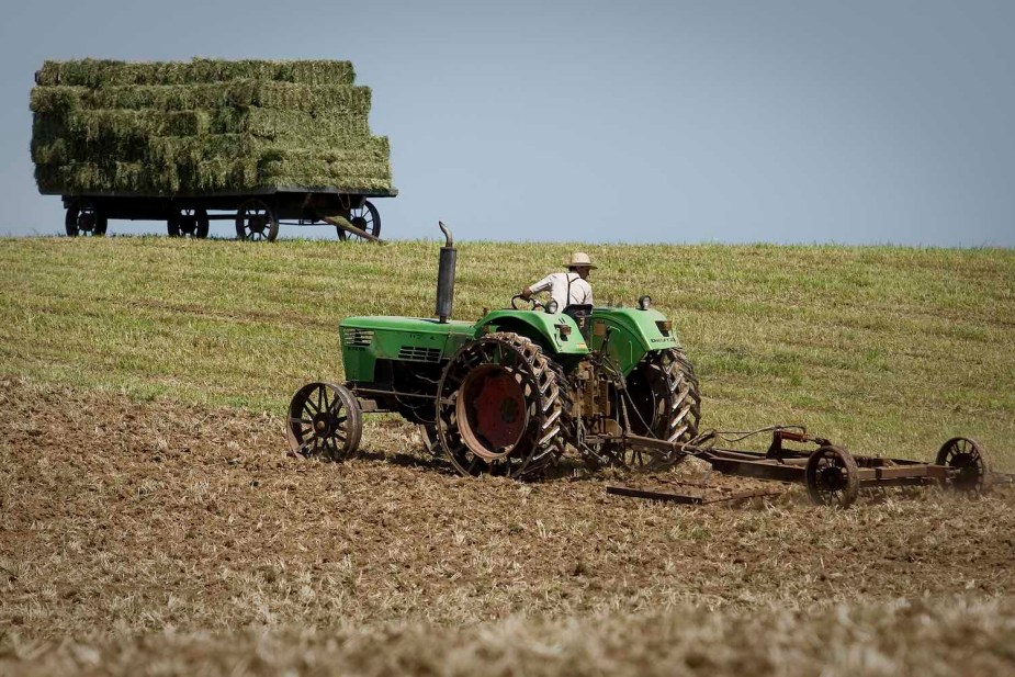 An Amish farmer rides his tractor with steel wheels through his field, a hay trailer parked in the background.