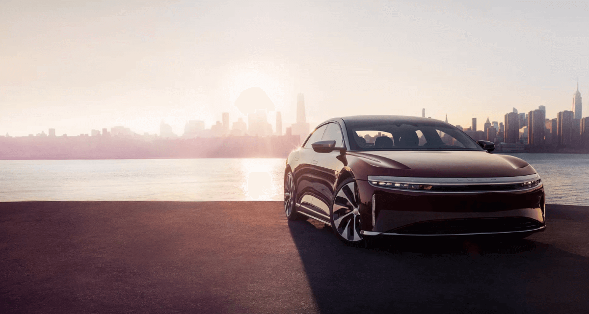 A Lucid Air all-electric luxury sedan model parked on a beach near a river and city skyline as the sun sets