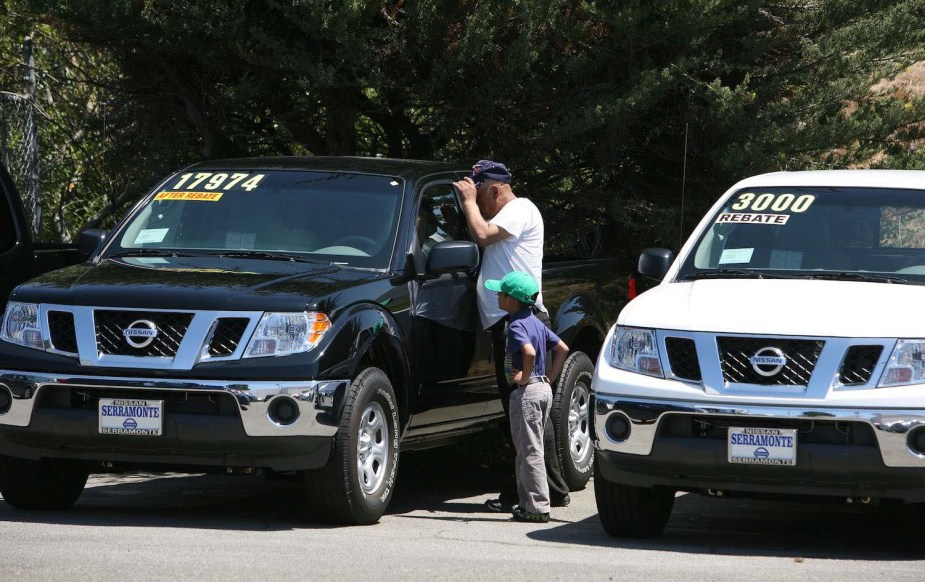 A man and a kid look in the window of a used Nissan pickup truck on a car lot.