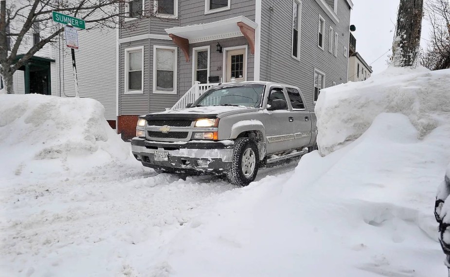 Silver diesel truck starting in the cold winter snow, a house visible in the background.