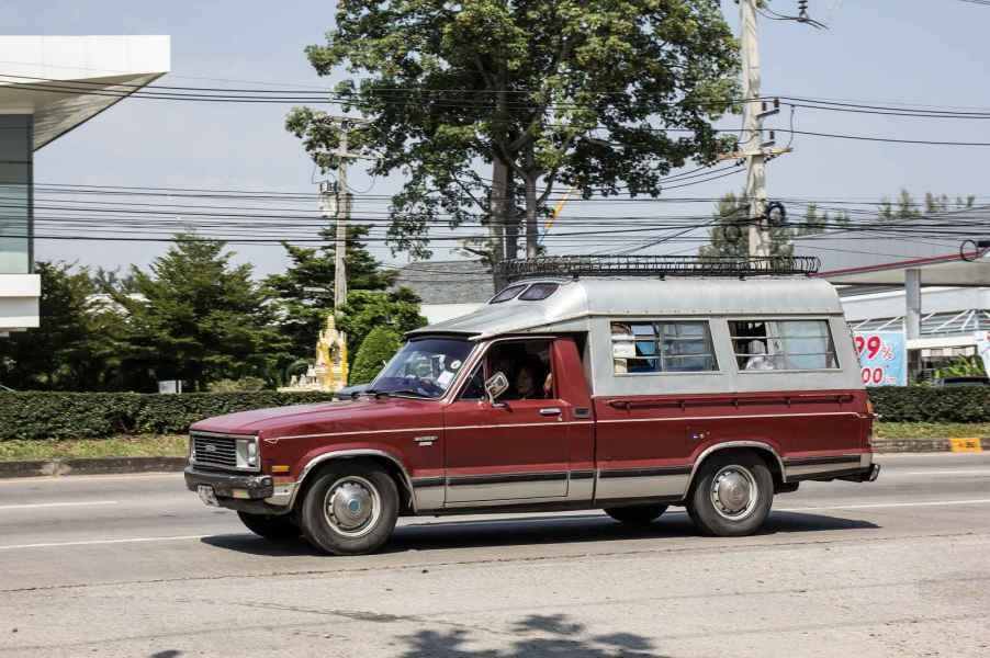 A burgundy colored Mazda B2200 compact pickup truck with an aluminum bed cap drives on the road in left profile view