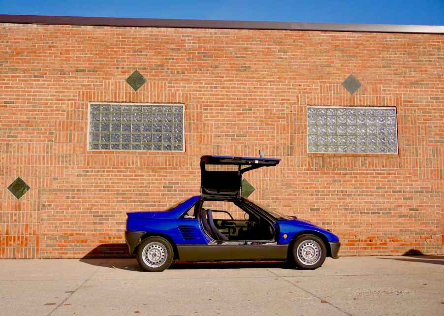 Blue Autozam AZ-1 sports car by Mazda with its gull-wing door open, a brick wall visible in the background