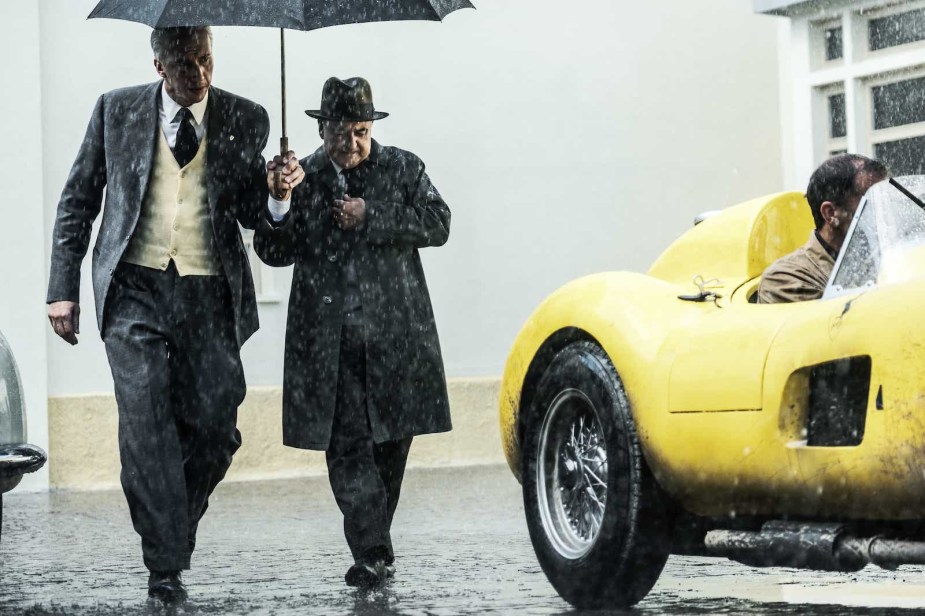 Suited men under an umbrella, standing by a yellow race car.