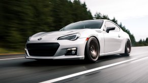 Silver Subaru BRZ sports car races along a road, trees visible in the background.