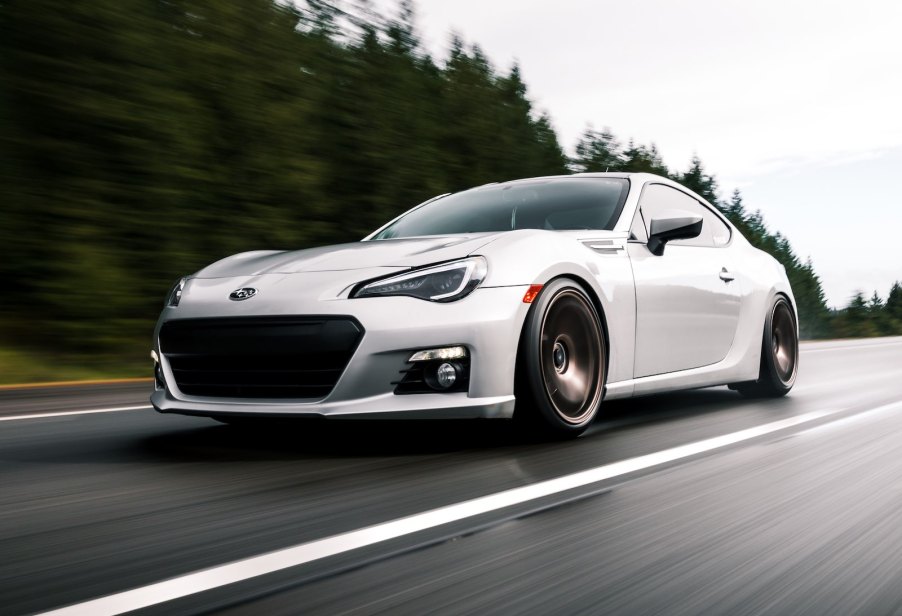 Silver Subaru BRZ sports car races along a road, trees visible in the background.