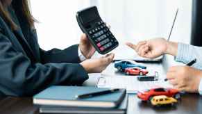 A woman holds a calculator up to a man sitting across from her with journals, toy cars, paperwork on the desk discussing car loan payments or trade-in