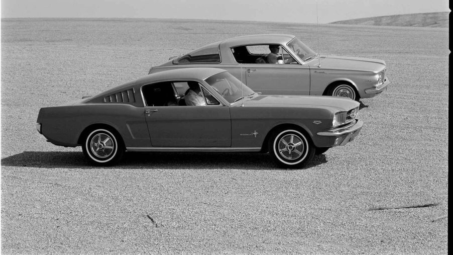 A 1965 Ford Mustang sits next to a 1965 Plymouth Barracuda in the desert the first pony cars black and white photo