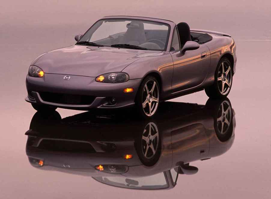 A grey 2004 Mazda MX-5 Miata shown parked on a reflective surface in left front angle view with yellow low beam lights on pink sunset colors