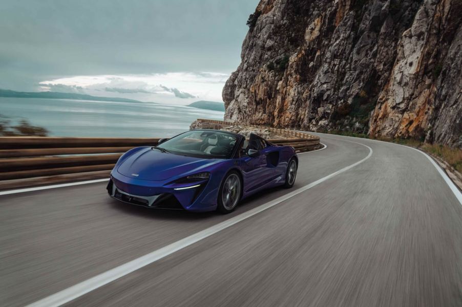 A blue 2025 McLaren Artura Spider cruises around a coastal road.