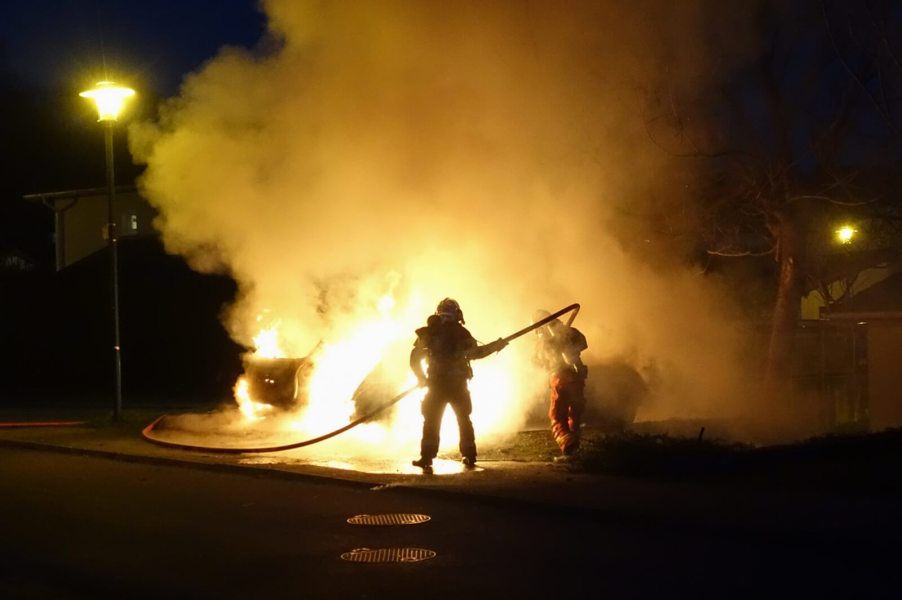 Firefighters battle a car fire with a vehicle like a Tesla or BMW.