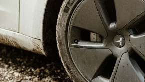 The rim and tire on a dirty white Tesla, parked on a dirt road.