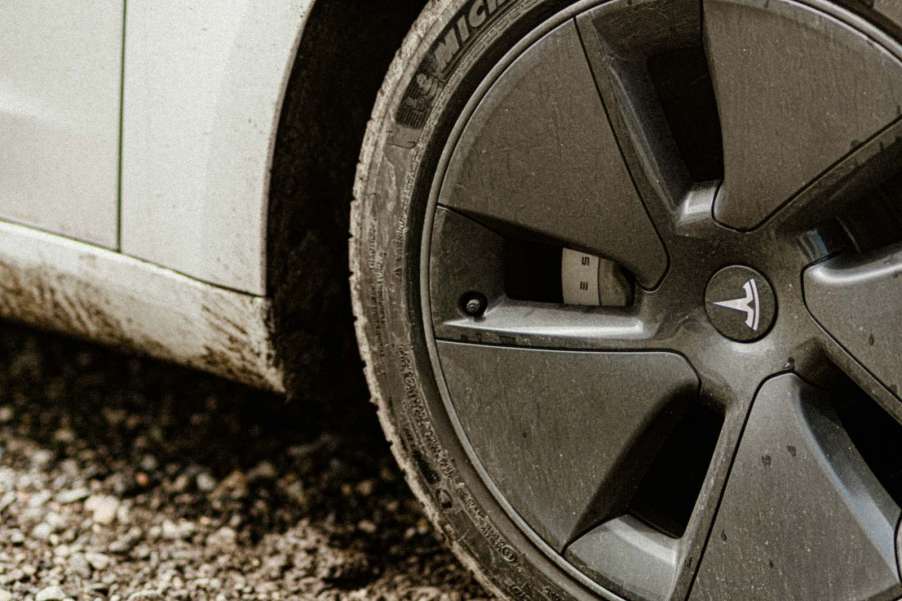 The rim and tire on a dirty white Tesla, parked on a dirt road.