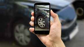 A hand holds up a cell phone taking a photo of a black car with right fender damage