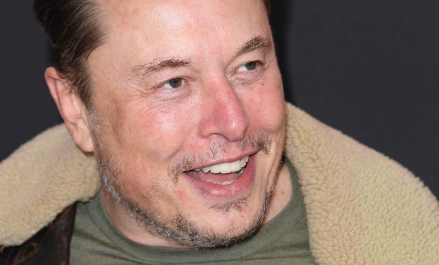 Tesla CEO Elon Musk shown smiling wide in close angle view Elon looking to his left slightly wearing a shearling-lined jacket