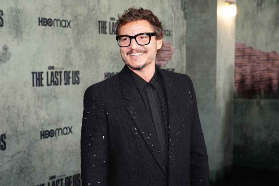 Marvel star Pedro Pascal at a media event wearing all black and black framed glasses with mustache and smiling