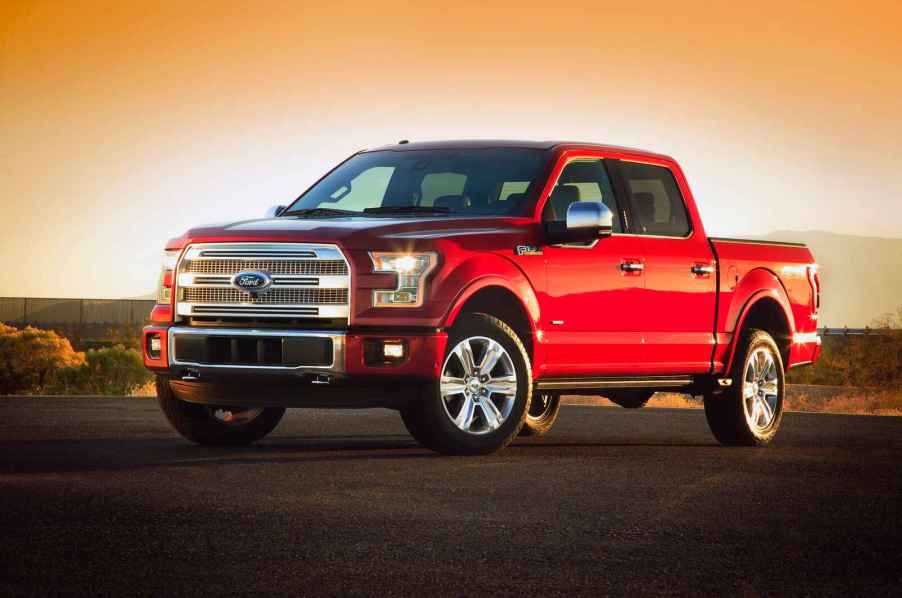 A 2015 Ford F-150 pickup truck parked in left front angle view front wheels turned in headlight glimmering sun setting behind parked on smooth black pavement